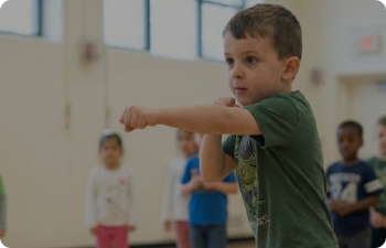 A boy in a green shirt punches the air as children watch from behind him in an indoor basketball court.