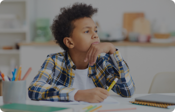 A boy sitting at a desk, looking to the right, hand cradling his chin, and a pencil grasped in hand.