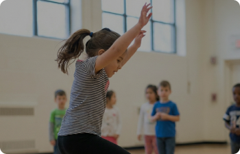 A girl with a dark-haired ponytail begins a cart-wheel in a basketball court with children watching behind her