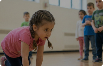 A girl in a pink shirt and with double dutch braided brown hair crawls on her hands and knees across the floor of an indoor basketball court as children behind her watch.