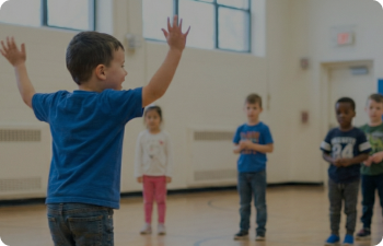 A boy in a blue shirt runs away from the camera with his hands raised in and indoor basketball court as children behind him watch.