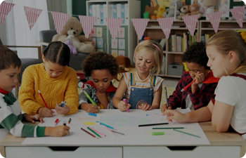 Six children doodling on a big white paper while sitting at a large desk in a classroom.